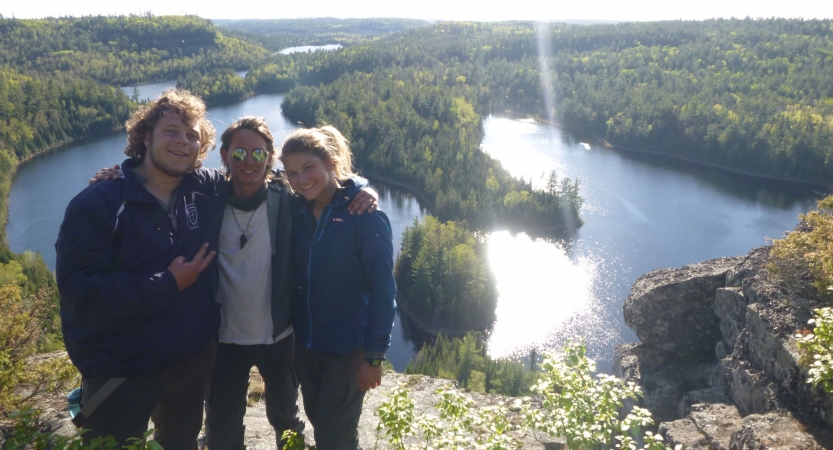 Three people pose for a photo with their arms around each other. They appear to be standing on a lookout, over a bending body of water and green woods.